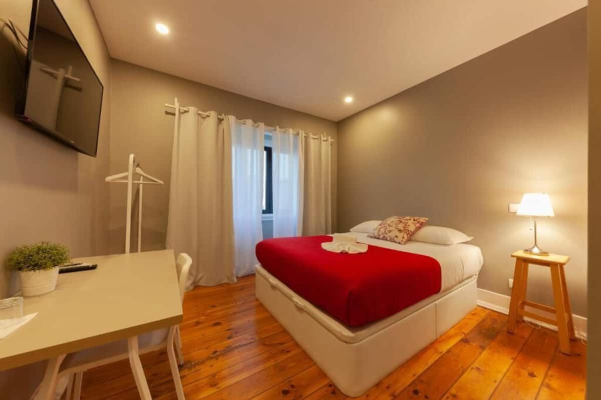 A bedroom with a bed, a desk, a chair, a TV mounted on the wall, and a window with curtains.