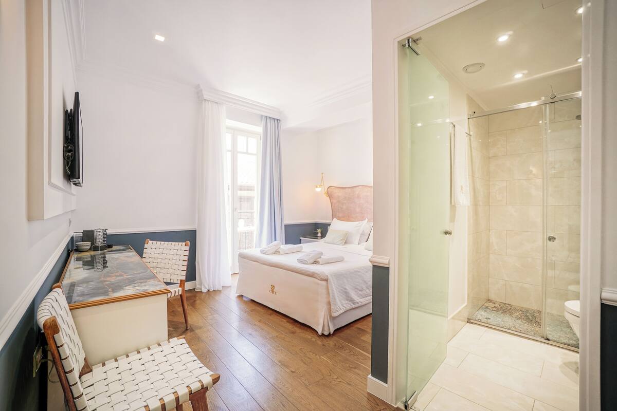 A modern hotel room with a bed, a seating area with a table and chairs, a glass-enclosed shower, and a view of the city.