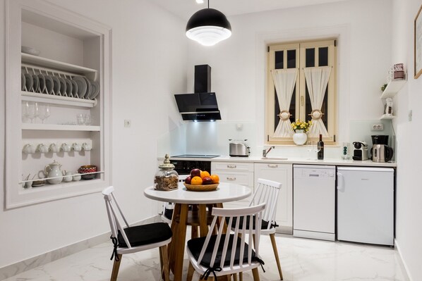 A modern kitchen with white cabinetry, a round dining table with two chairs, a built-in oven, and a coffee machine.