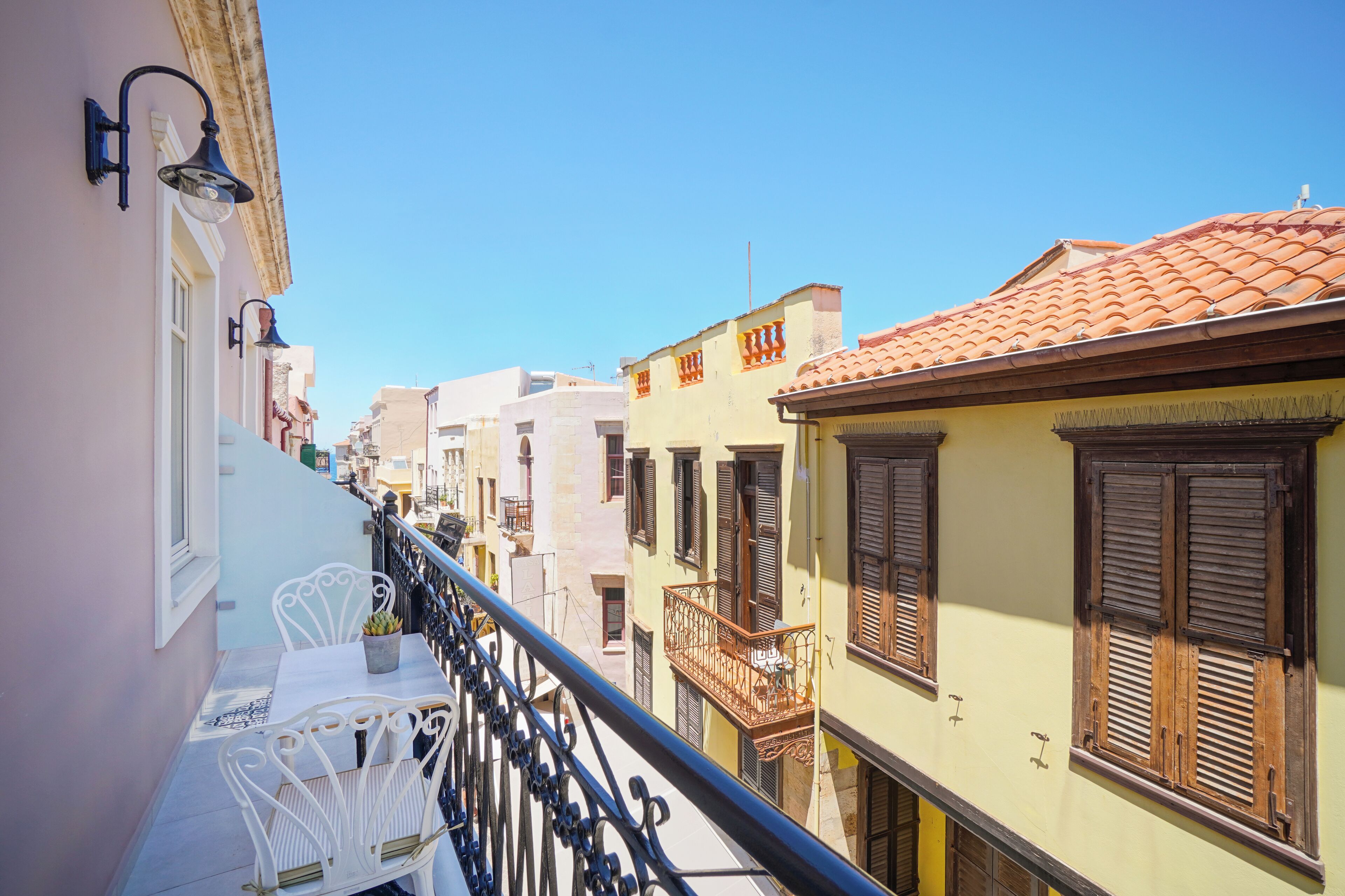 A balcony with white metal chairs and a small table, overlooking a narrow street with colorful buildings and a clear blue sky.