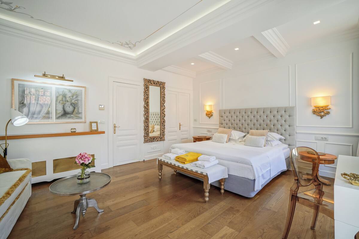 A hotel room with a large bed, a bedside table, a chair, a small round table, and a painting on the wall.