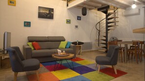 Una sala de estar con un sofá, dos sillones, una mesa de centro, un tapete colorido, una escalera en espiral y un comedor con sillas de madera y una mesa.