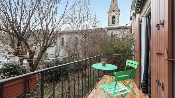 Un balcon avec une table et des chaises vertes, donnant sur une cour intérieure où l’on aperçoit, au loin, le clocher d’une église.