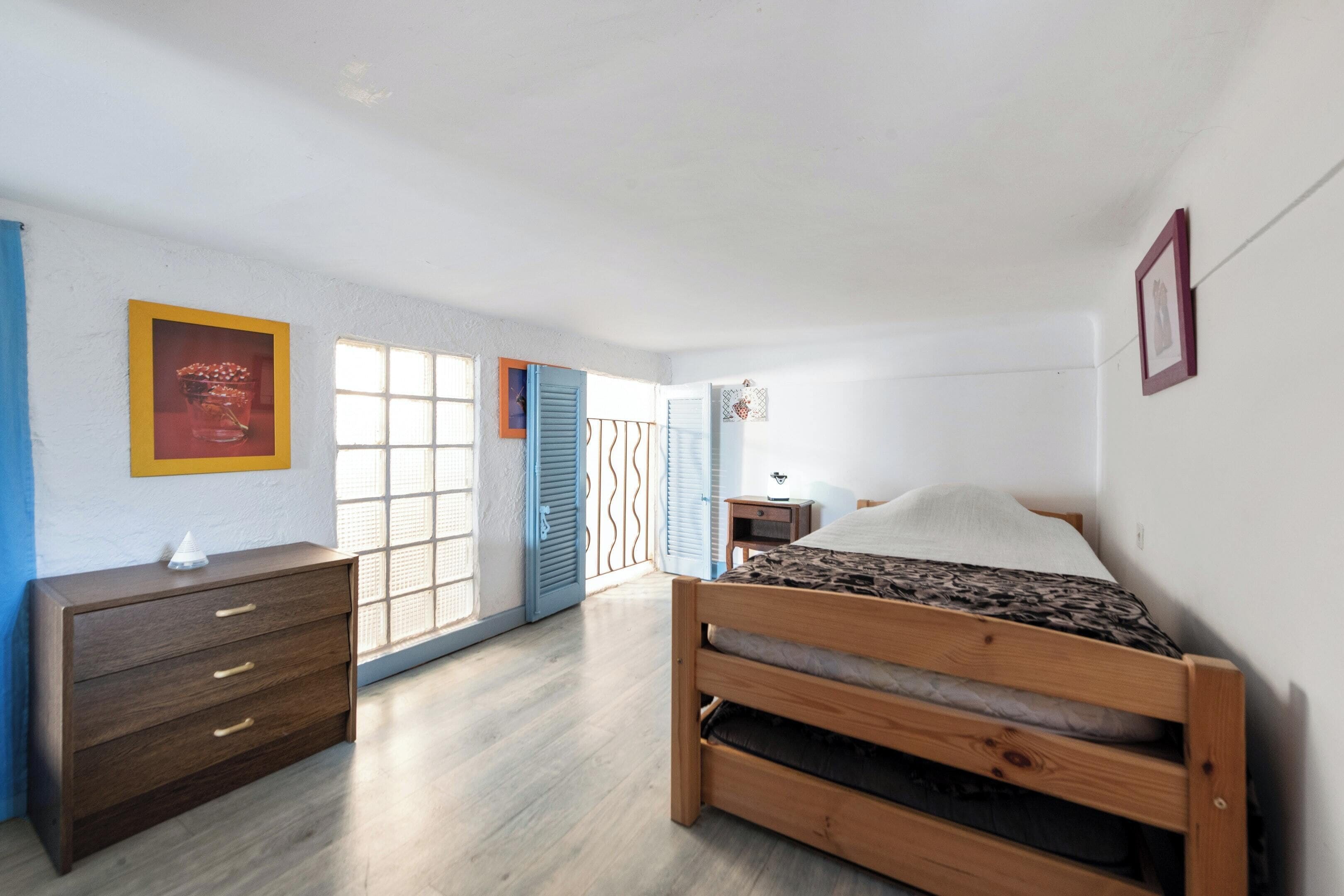 Ein Schlafzimmer mit einem hölzernen Bett, einem Schminktisch, einem Fenster mit blauen Fensterläden und einer Tür zu einem anderen Raum.