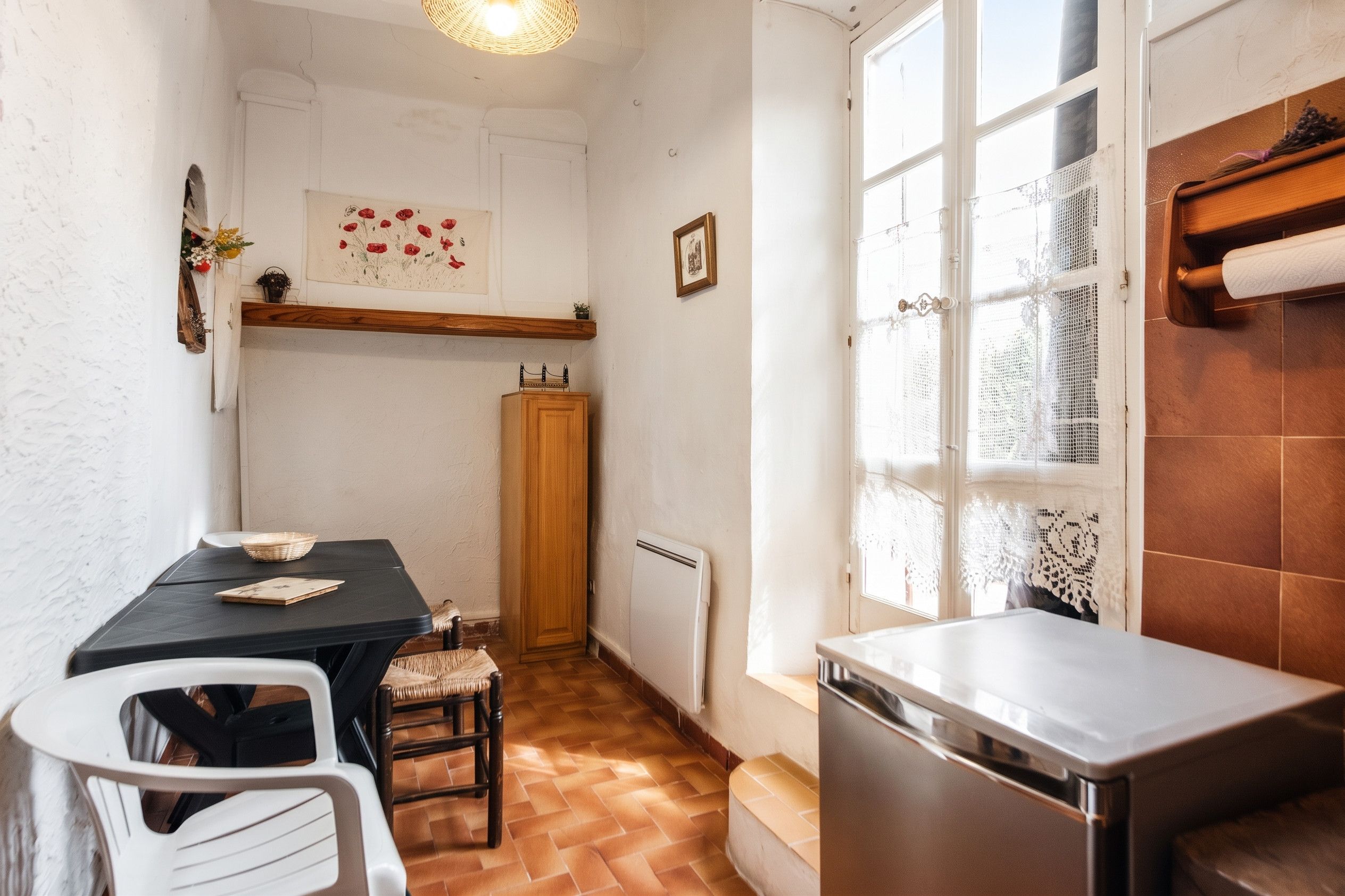 A compact kitchen with a dining table, chairs, a refrigerator, and a window with lace curtains.