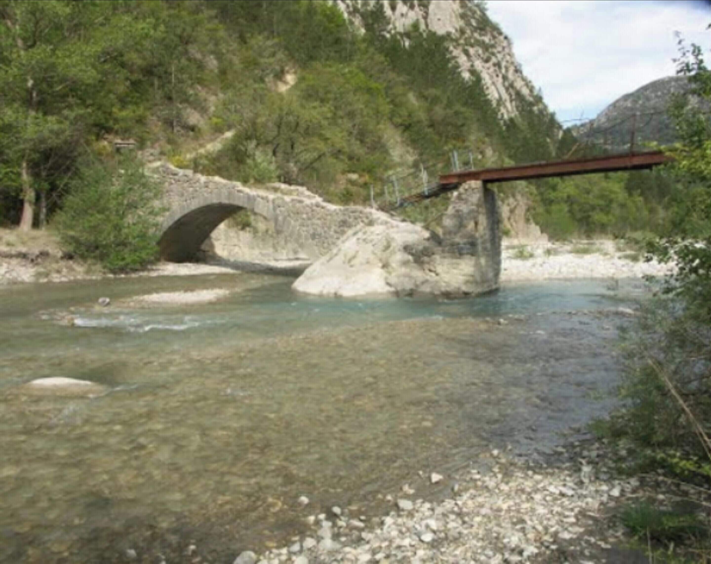 Een stenen brug over een heldere rivier met rotsachtige oevers en begroeiing op de achtergrond.