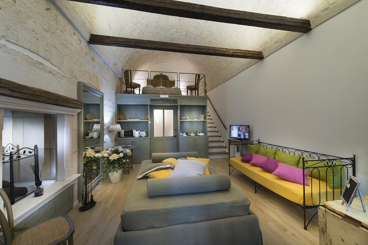 A cozy loft-style living space with a loft bed, a sofa with colorful cushions, a dining area with a table and chairs, and a kitchen area with shelves and appliances.