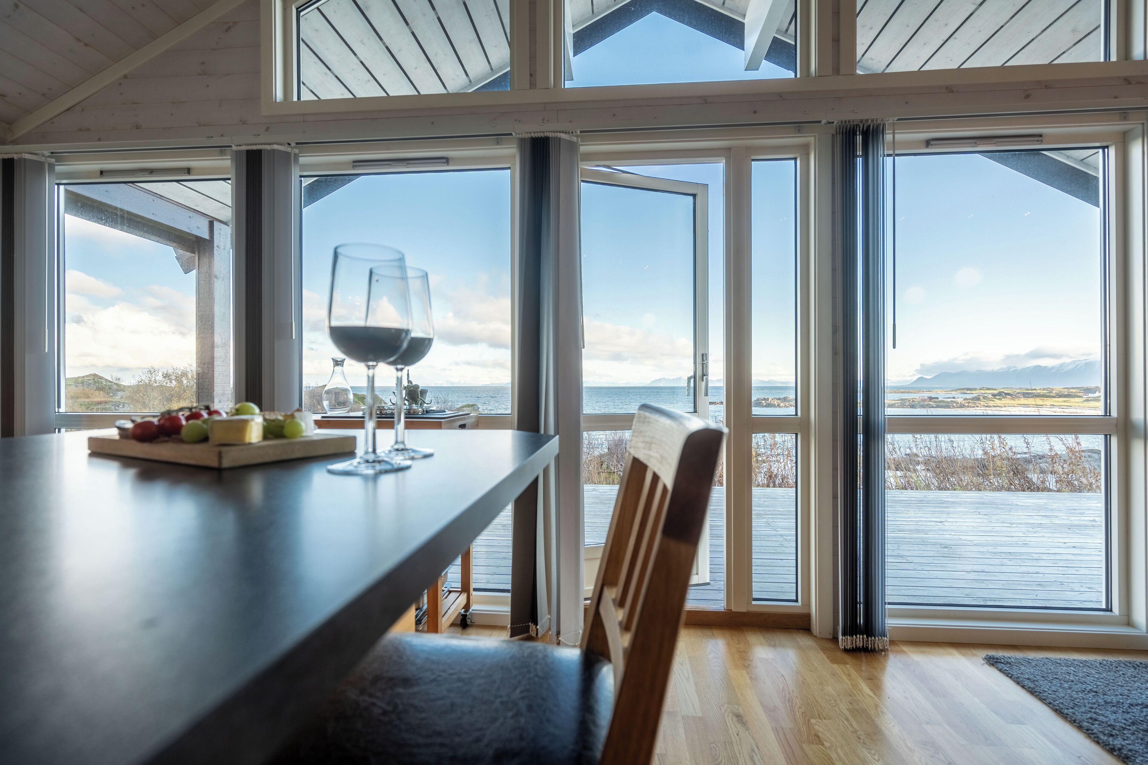 A dining area with a table set for two, a glass of red wine, and a fruit platter. Large windows offer a view of the water and mountains.