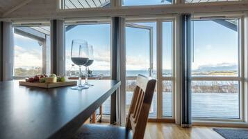 A dining area with a table set for two, a glass of red wine, and a fruit platter. Large windows offer a view of the water and mountains.