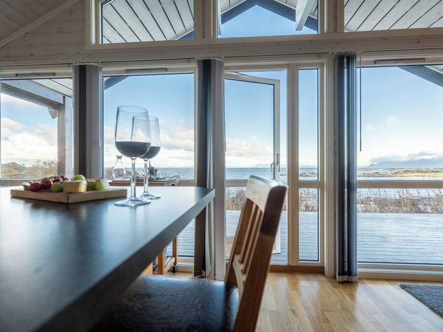 A dining area with a table set for two, a glass of red wine, and a fruit platter. Large windows offer a view of the water and mountains.