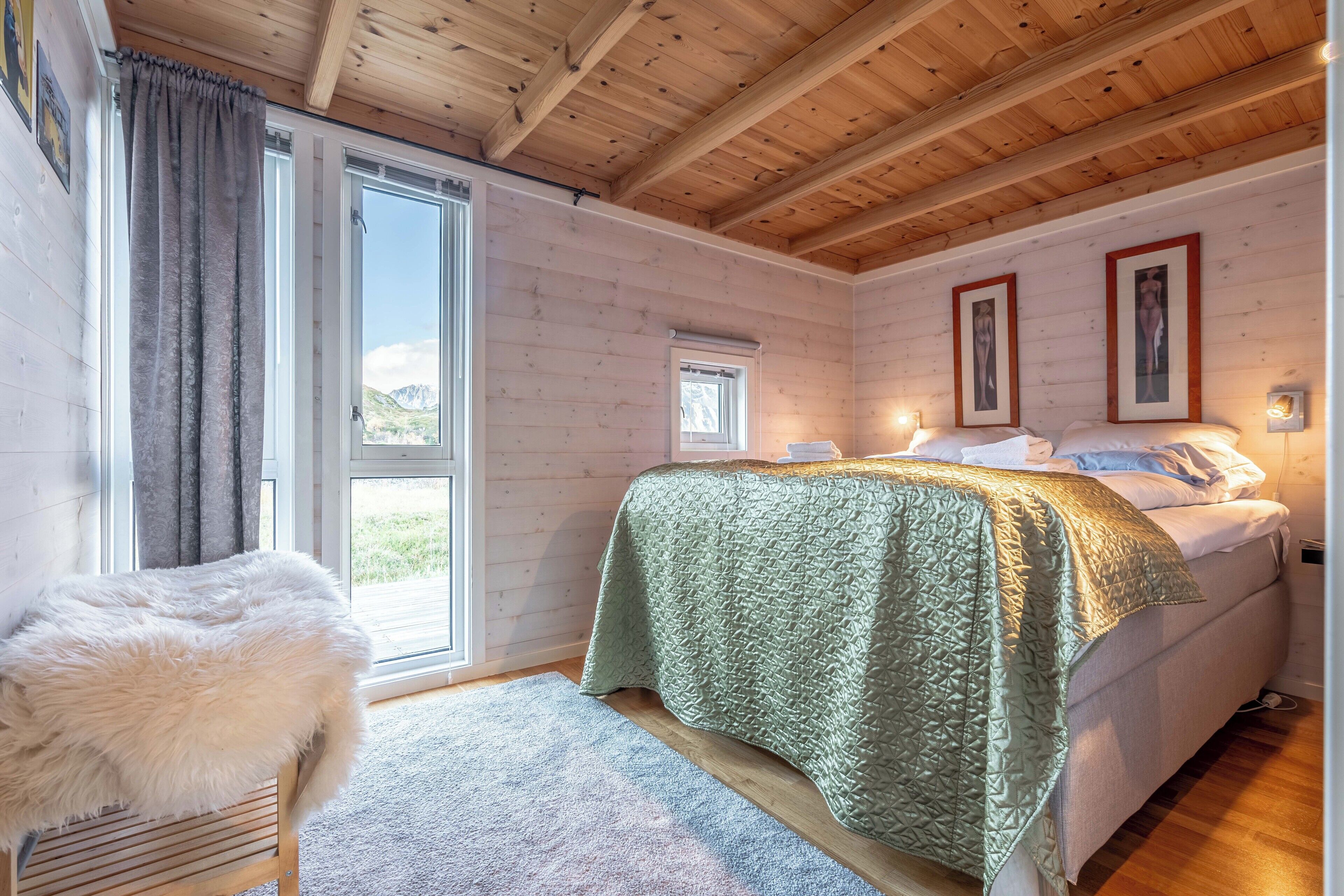 Une chambre confortable avec un plafond en bois, une grande fenêtre, un lit recouvert d’une couverture verte et un fauteuil blanc moelleux.