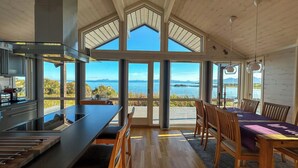 Cocina moderna con una isla grande, mesa de comedor de madera con sillas y ventanas amplias que ofrecen vistas al mar y a las montañas.