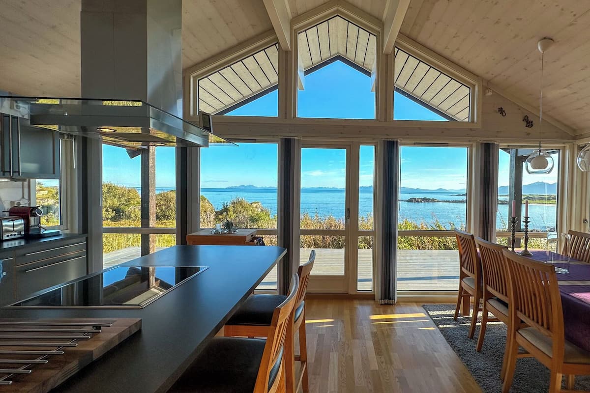 A modern kitchen with a large island, wooden dining table with chairs, and large windows offering a view of the sea and mountains.