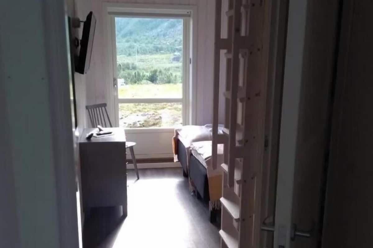 A narrow hallway leading to a bedroom with a bed, a desk, and a chair. A window offers a view of greenery outside.