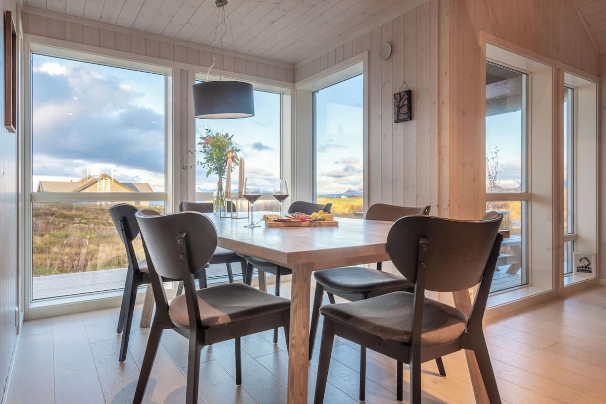 A dining area with a wooden table and chairs, large windows with a view of the landscape, and a vase with flowers on the table.