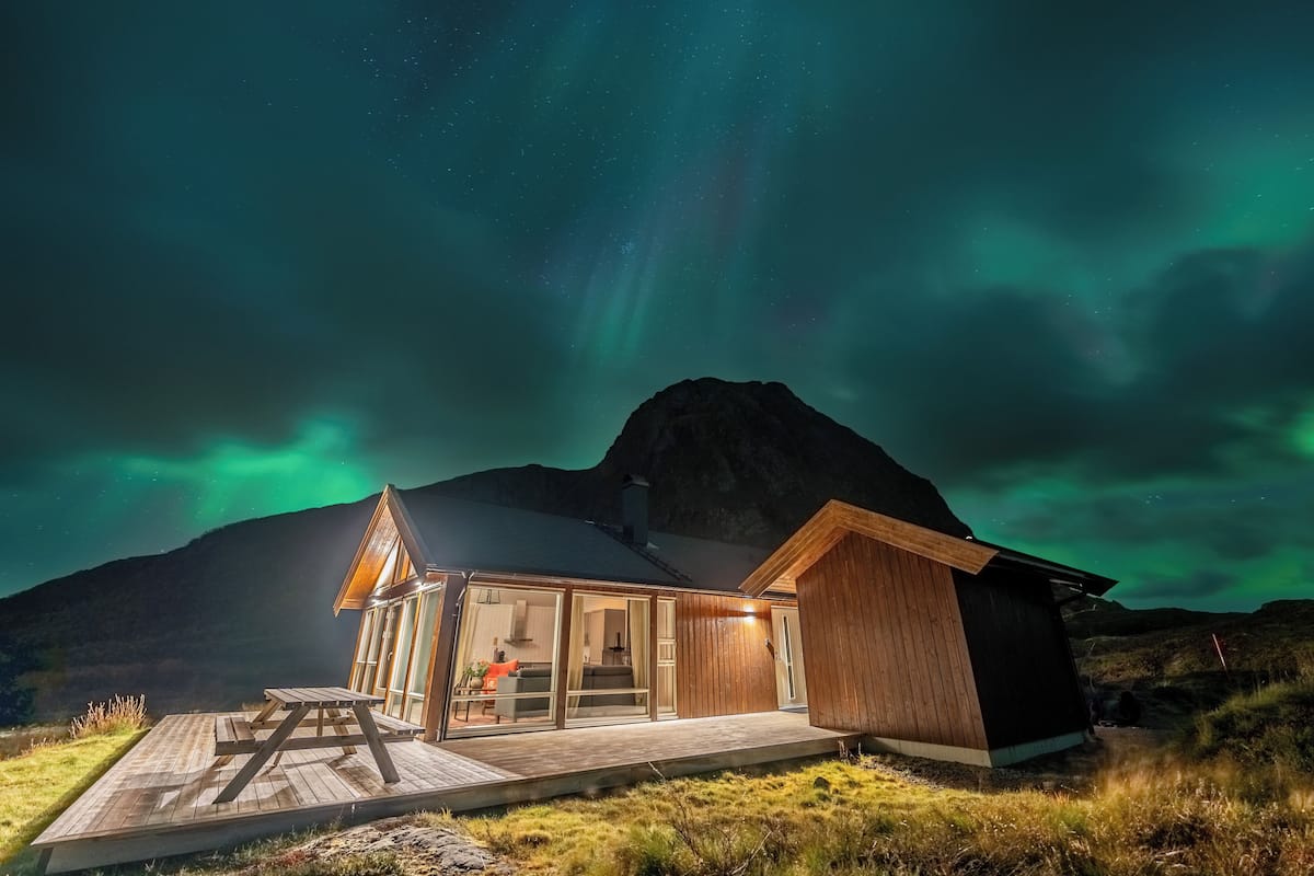 A wooden cabin with a glass door entrance, a deck, and a mountain backdrop.