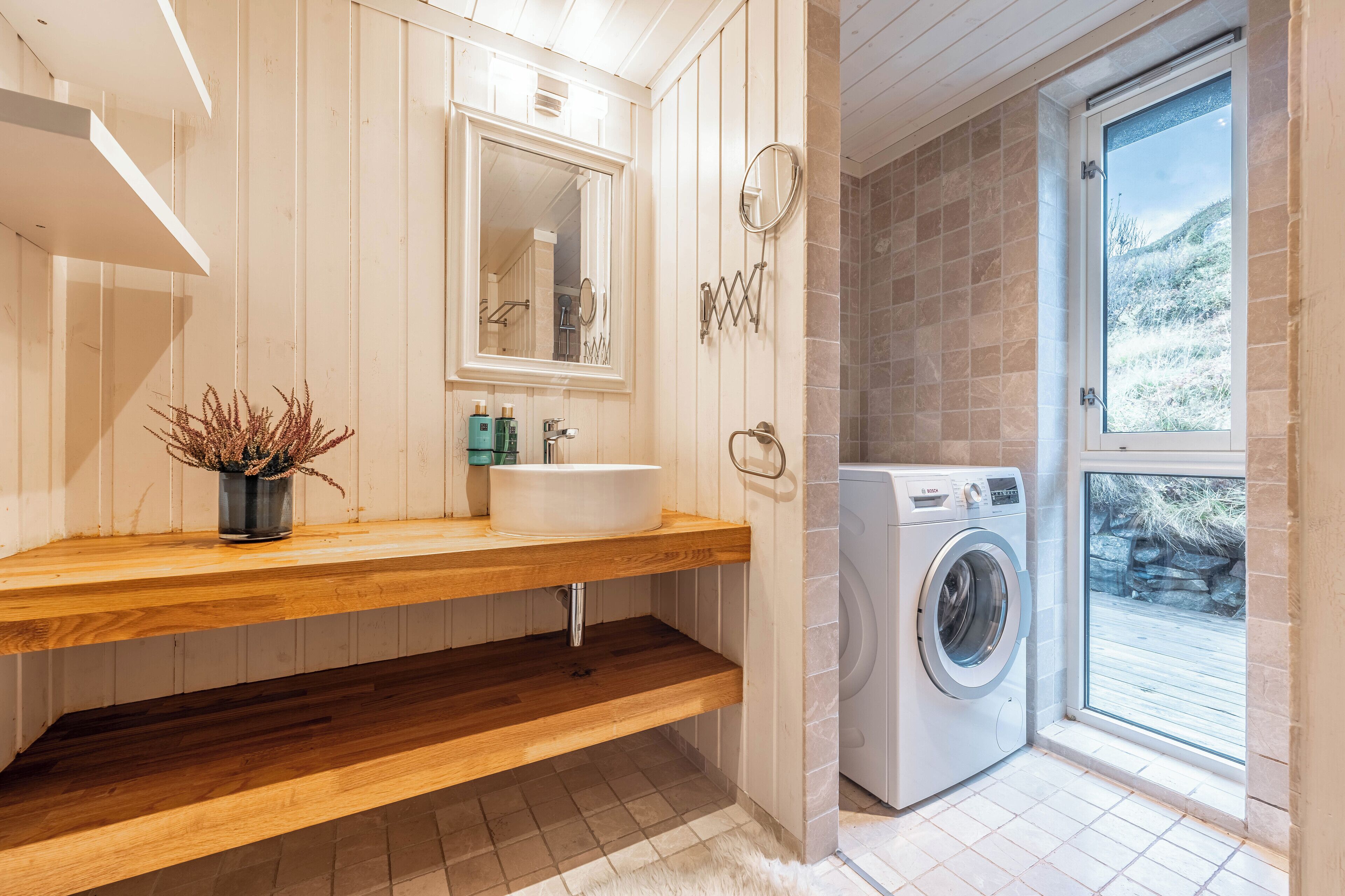 A modern bathroom with a wooden vanity, a large mirror, a built-in washing machine, and a view of a mountainous landscape through a glass door.