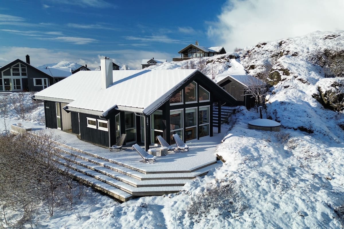 A modern chalet with a snow-covered roof, large windows, and a stone staircase leading to the entrance.