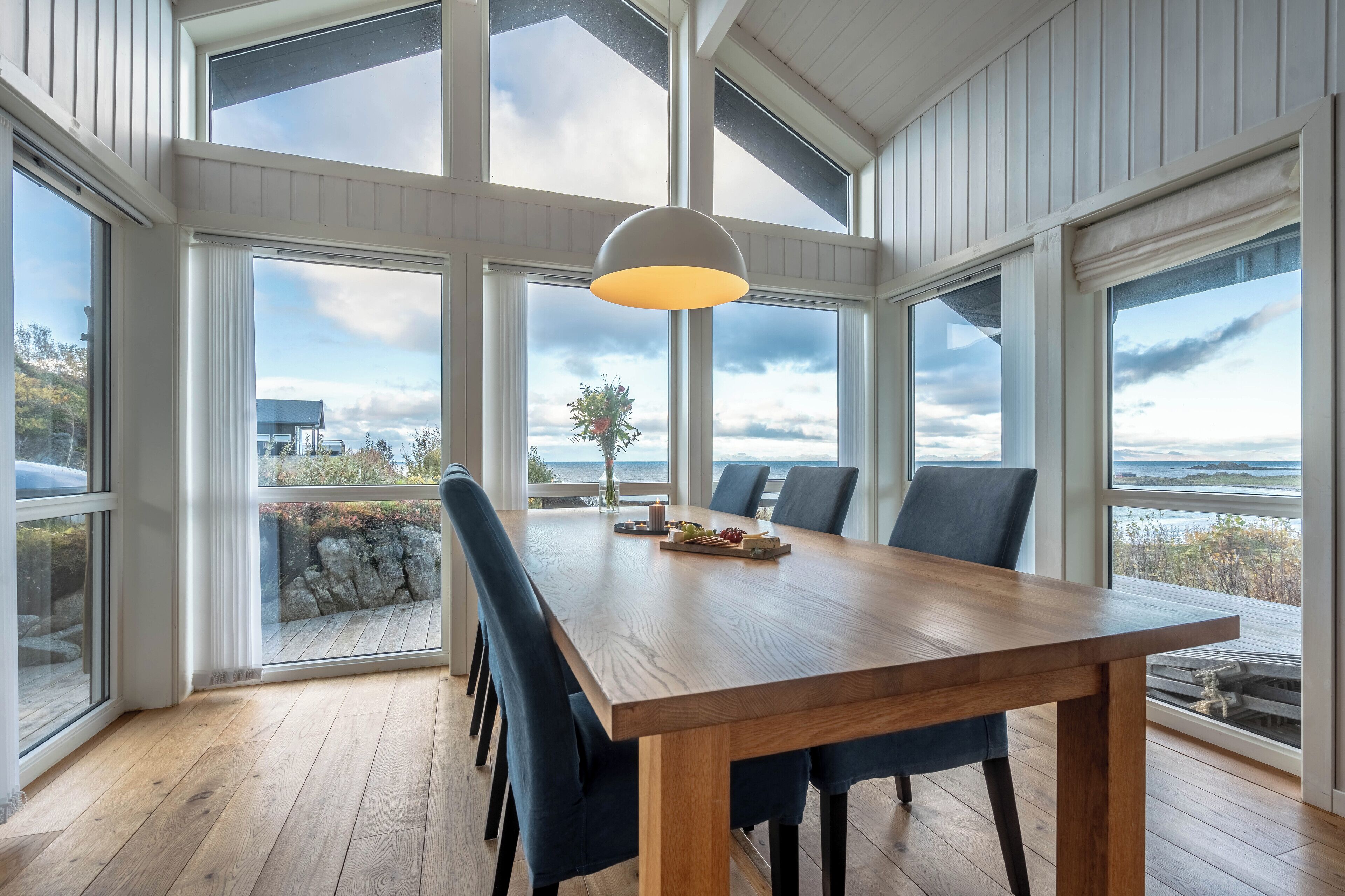 A spacious dining area with a wooden table, blue chairs, and a large window offering a view of the ocean and a rocky shoreline.
