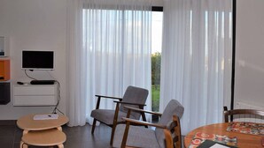 A modern room with a flat-screen TV, a wooden chair, a small round table, and a large window with sheer curtains. - Modern Single-Level House with WiFi, Terrace, and Parking in Trégastel (Trégastel)