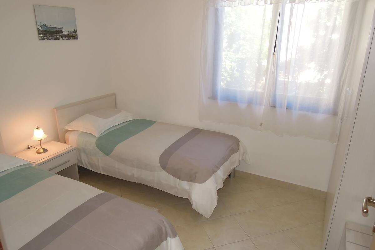 A bedroom with two beds, a nightstand with a lamp, a window with sheer curtains, and a painting on the wall.
