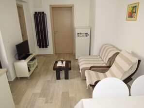 A living room with a sofa, a coffee table, a TV on a stand, a refrigerator, and a door with a towel rack. - Apartmani Simicev (Zadar)
