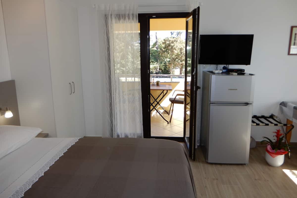 A bedroom with a bed, a television on a stand, a refrigerator, and a view of an outdoor patio with a table and chairs.