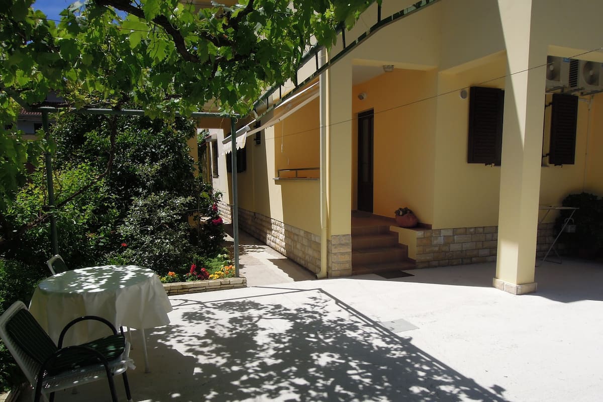A covered outdoor patio with a table and chairs, a staircase leading to a door, and a garden with various plants.
