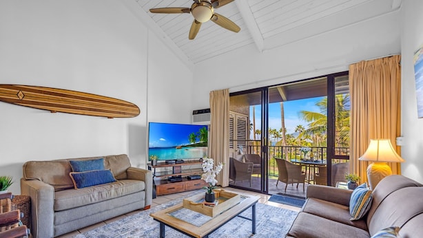 Ein Wohnzimmer mit Sofa, Couchtisch und Fernseher, mit Blick auf Palmen und einen Balkon.
