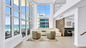 A spacious living area with large windows, a white sofa, two armchairs, a coffee table, and a bookshelf.