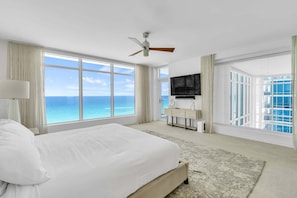 Hotelkamer met een bed, een plafondventilator, een televisie, een groot raam met uitzicht op de oceaan en een klein bureau met stoel.