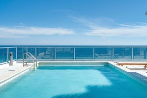 Une piscine aux eaux d’un bleu limpide, bordée d’une rambarde en métal, avec une terrasse en bois et une vue sur l’océan.
