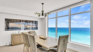 A dining area with a large window overlooking the ocean, a chandelier, and a modern kitchen visible through the glass wall.