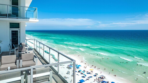 A balcony with outdoor seating overlooking a beach with clear turquoise water and a white sandy shore.