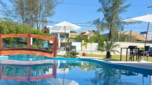 Piscina azul límpida com um deck de madeira e espreguiçadeiras, rodeada por árvores e edifícios.
