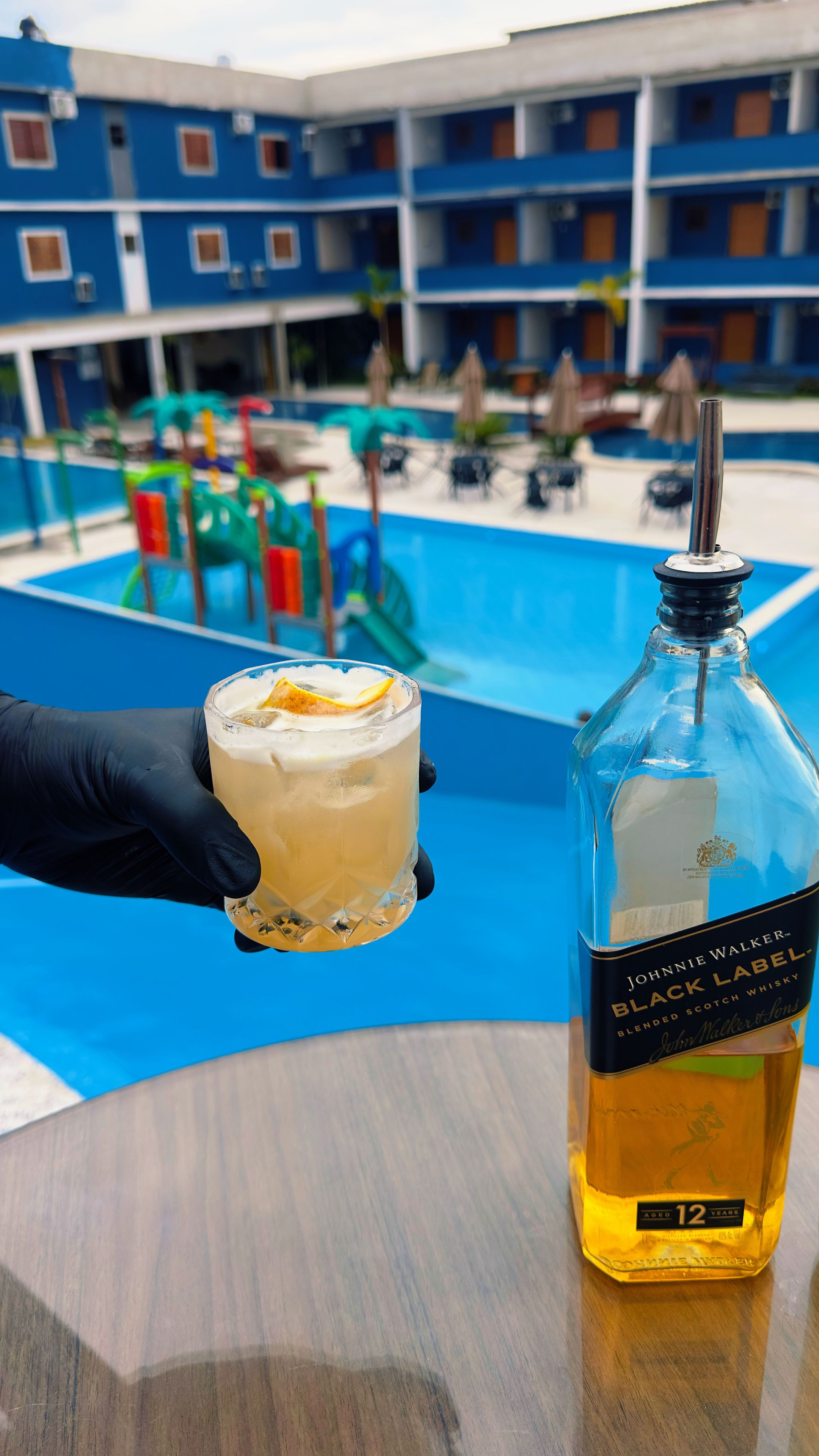 Una mano sosteniendo un vaso con una bebida, una botella de whisky Johnnie Walker Black Label, y un área de piscina con sombrillas y sillas de descanso al fondo.