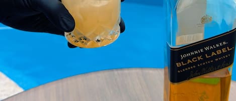 A hand holding a glass with a drink, a bottle of Johnnie Walker Black Label whisky, and a pool area with umbrellas and lounge chairs in the background.