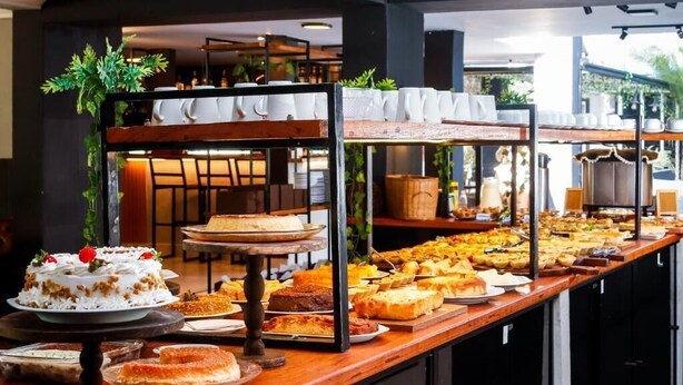Buffet com diversos pratos e sobremesas, incluindo bolos e doces, expostos em um balcão de madeira comprido.