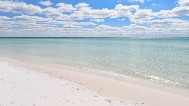 Een strand met helder turquoise water en wit zand, onder een blauwe lucht met enkele wolken.