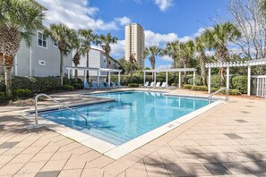 Una piscina con sillones de descanso y palmeras, rodeada por un edificio y una estructura alta al fondo.