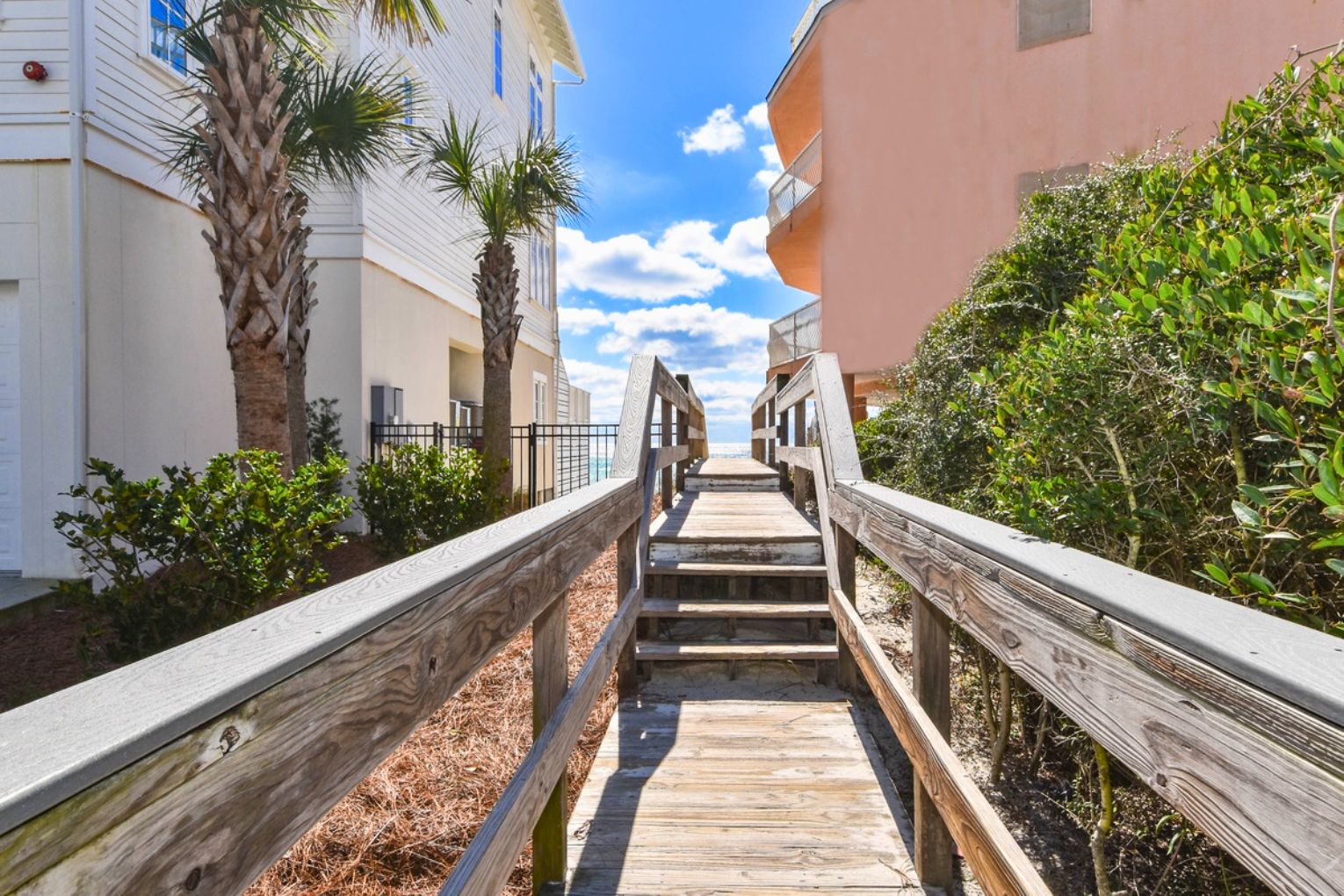 A wooden boardwalk leading to a building with a white facade and a pink building on the right.
