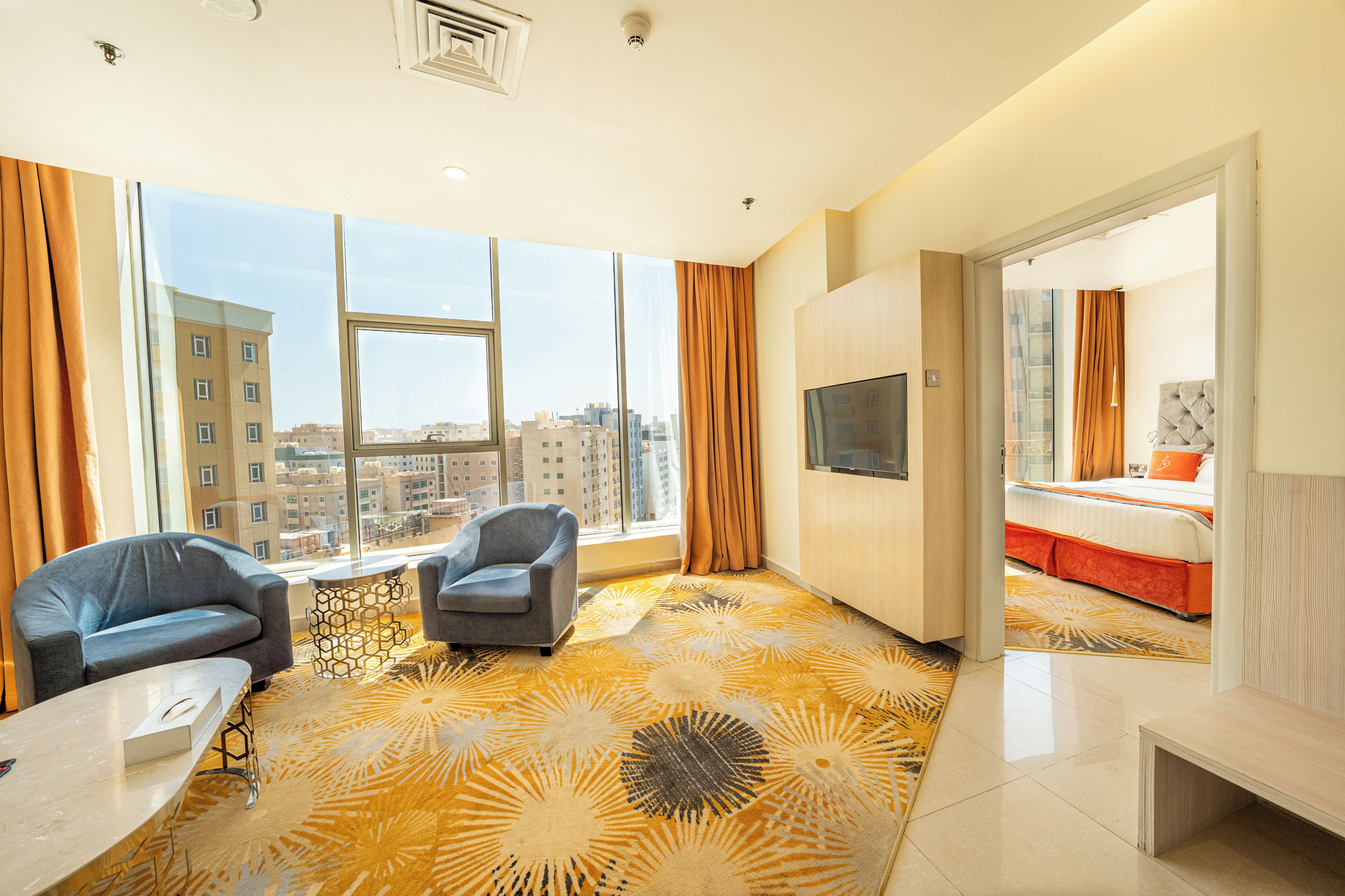 A hotel room with a large window, two armchairs, a bed with orange bedding, a TV, and a patterned carpet.