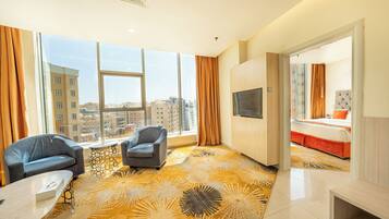 A hotel room with a large window, two armchairs, a bed with orange bedding, a TV, and a patterned carpet.