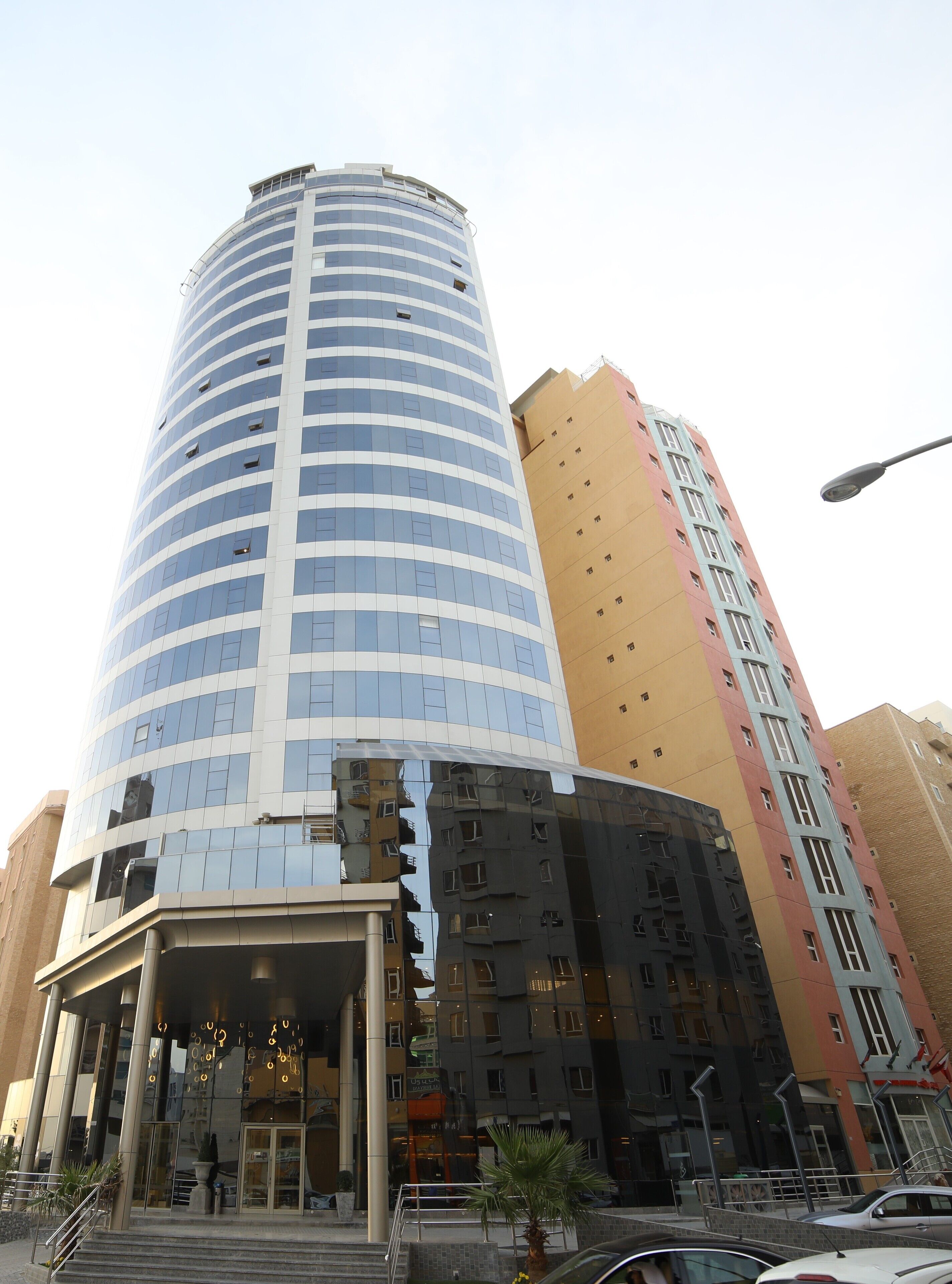 Un hotel moderno con fachada curva, amplias ventanas de vidrio y una entrada destacada con una palmera y zona de estar al aire libre.