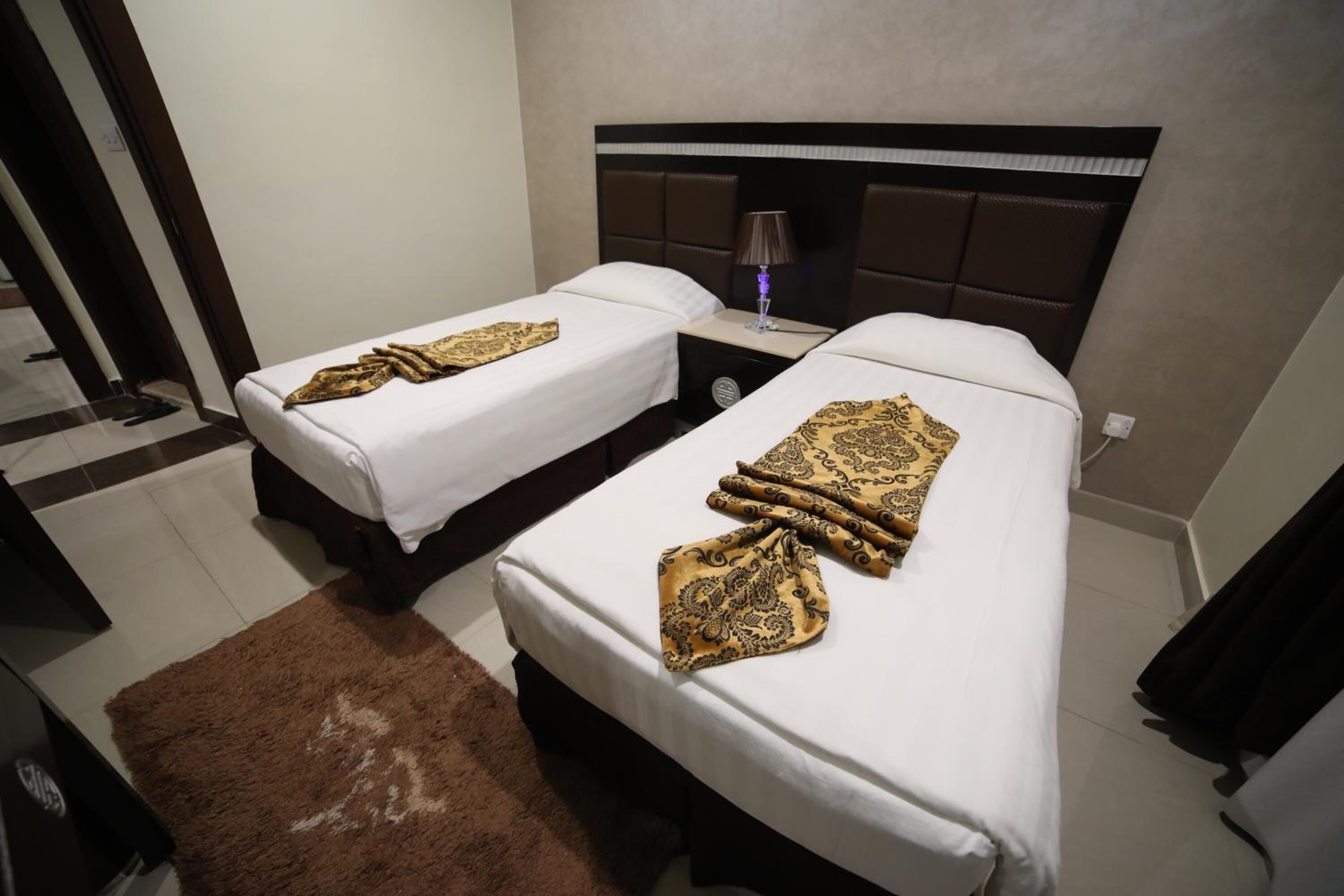 A hotel room with two single beds, a brown rug, a nightstand with a lamp, and a dark headboard.