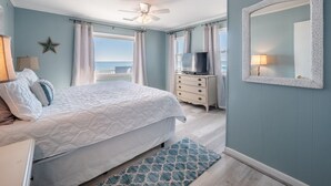 A bedroom with a bed, a dresser, a television, a ceiling fan, and a large mirror. - The Shark and the Seahorse | Ocean Views + Pet-Friendly (Surf City)