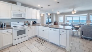 A modern kitchen with white cabinetry, a built-in oven, and a dishwasher. There is a large island with a sink and a dining area with a view of the ocean. - The Shark and the Seahorse | Ocean Views + Pet-Friendly (Surf City)