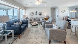 A spacious living room with a large window overlooking the ocean, a ceiling fan, and a dining area with a view. - The Shark and the Seahorse | Ocean Views + Pet-Friendly (Surf City)