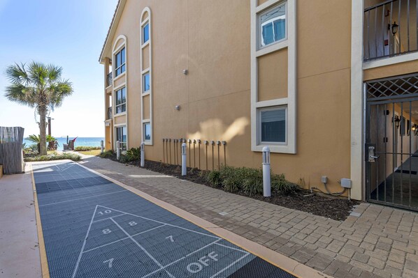 A building with a paved area marked for shuffleboard, a palm tree, and a view of the sea.