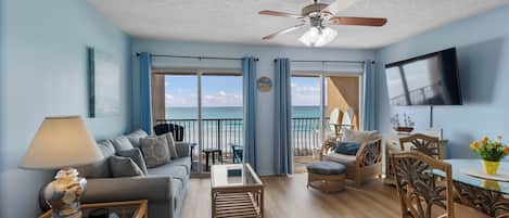A spacious living room with a large window overlooking the beach, a ceiling fan, and a flat-screen TV mounted on the wall.